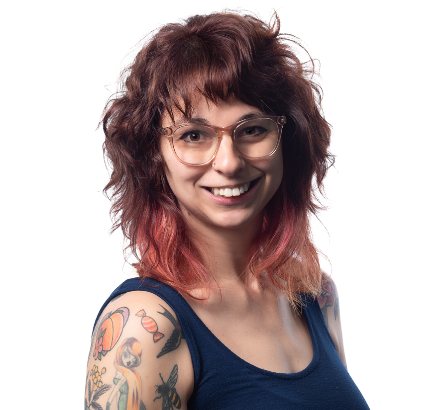 Emma Henningson, a womanwith shoulder length brown and pink hair smiles towards the camera. She is wearing glasses and has visible tattoos on her arm.