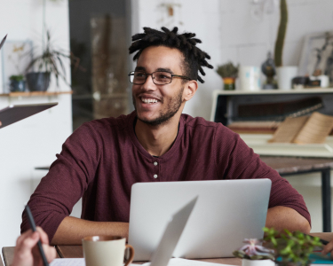 Man with dreadlocks and glasses sitting in front of laptop. Smiling off to the side.
