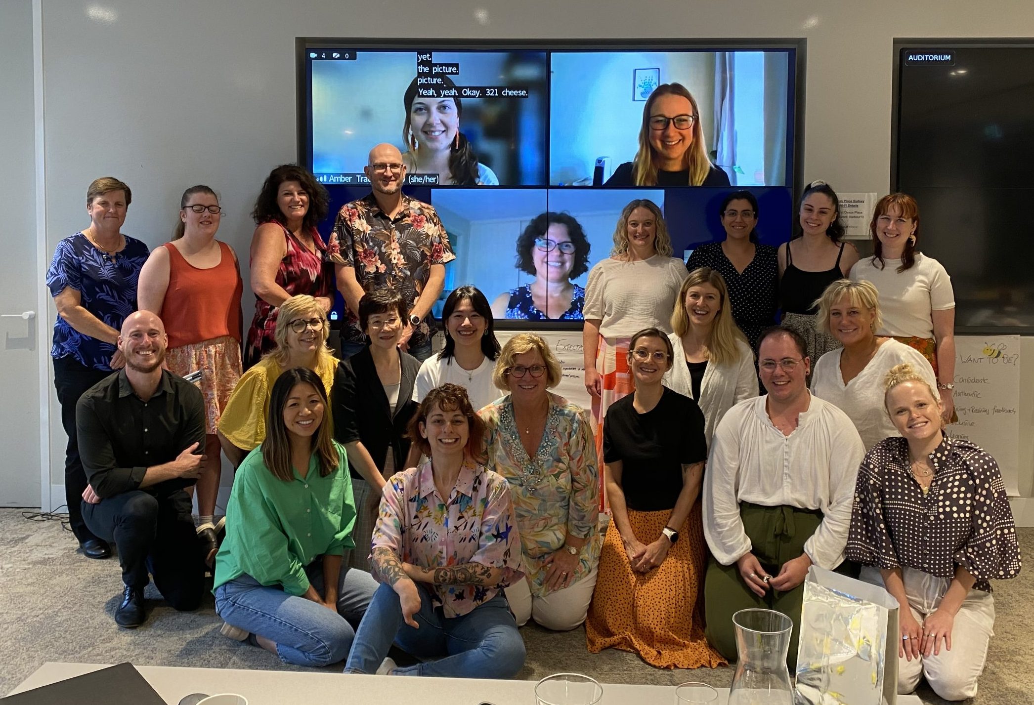 Australian Network on Disability team huddled together smiling. Three members of the team have joined virtually and are projected onto a screen behind the team.