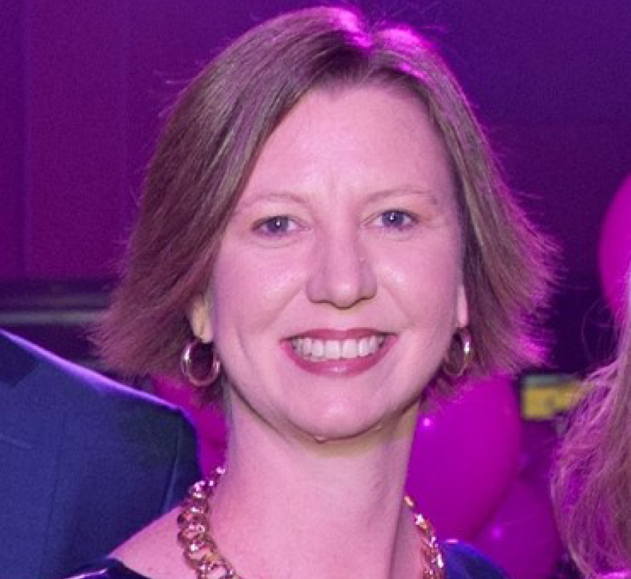 Picture of Karen Grumley, smiling towards the camera. She has chin length, fair brown hair and a large necklace on. The background is purple.